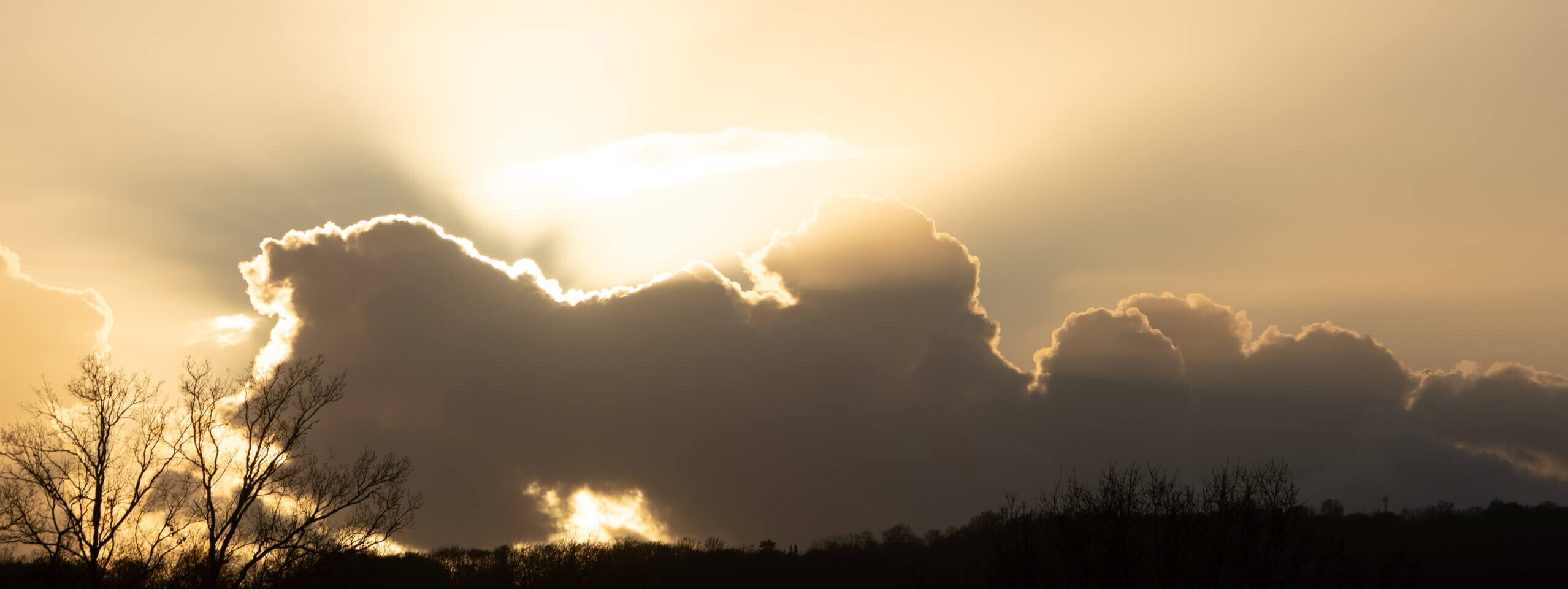Die Sonne strahlt durch eine Wolkenformation, die in sanften Gelb- und Grautönen leuchtet, während der Himmel sich aufhellt.